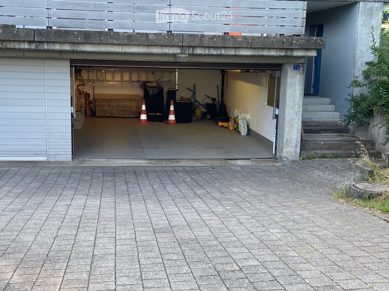 Garage with concrete floor, storage items, and traffic cones inside. Steps lead up to a raised ground floor area with a balcony above.