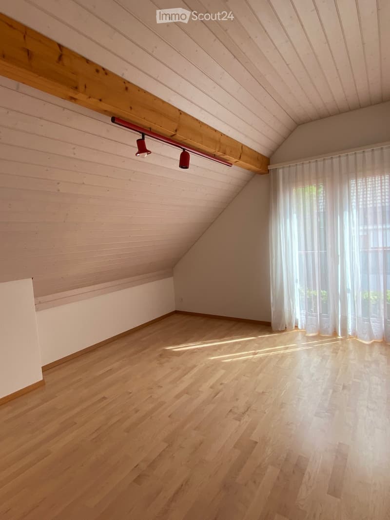 Empty attic room with wooden floors, white walls, and a slanted wooden ceiling. Natural light filters through sheer white curtains covering a window.