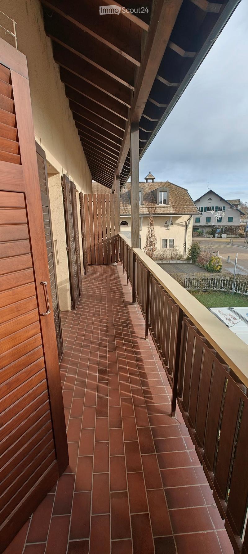 Balcony with terracotta tiled floor, wooden shutters, and a wooden railing, leading to a gated area with a view of a residential neighborhood.