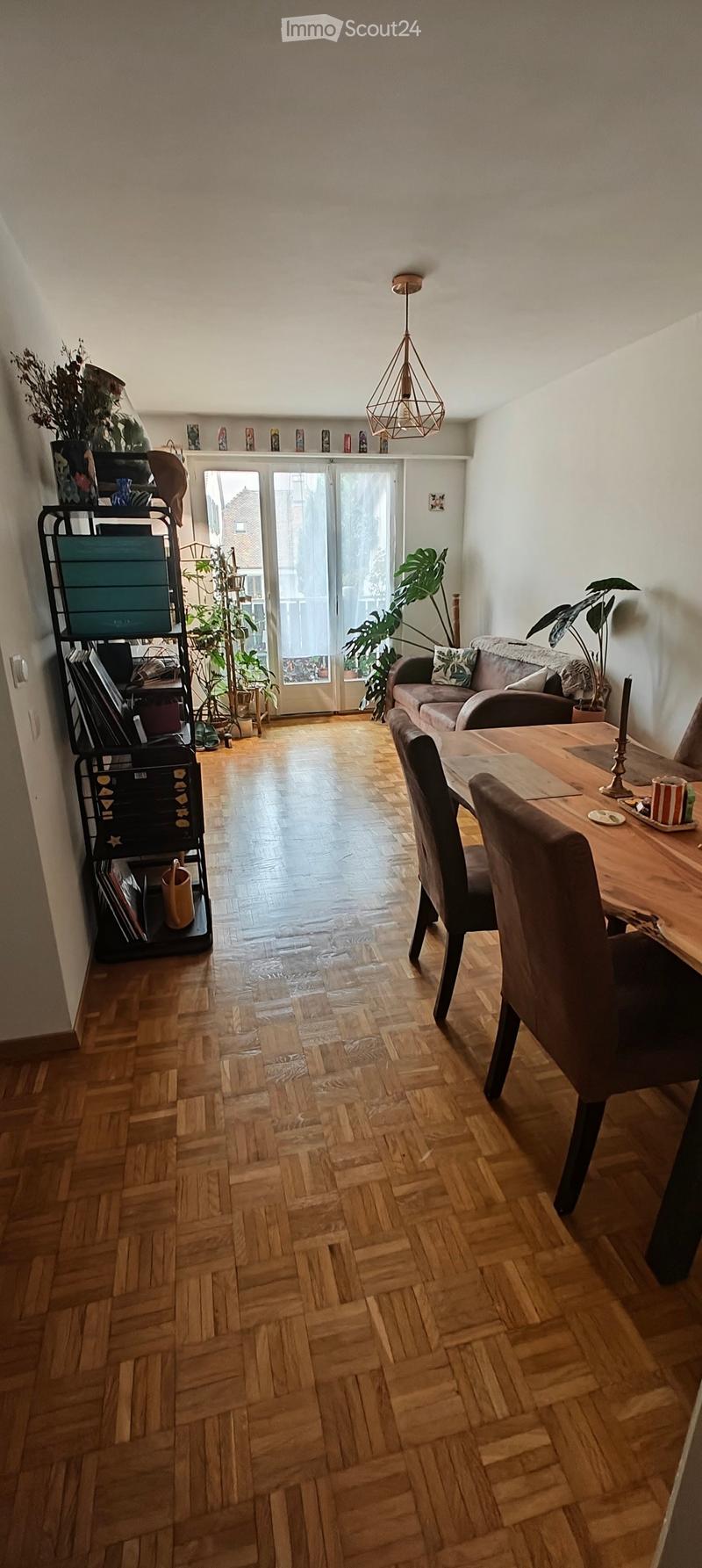 Living and dining area with wooden herringbone flooring, a grey sofa, a wooden dining table with chairs, a metal shelving unit, and several potted plants. A large window with a view leads to a balcony.