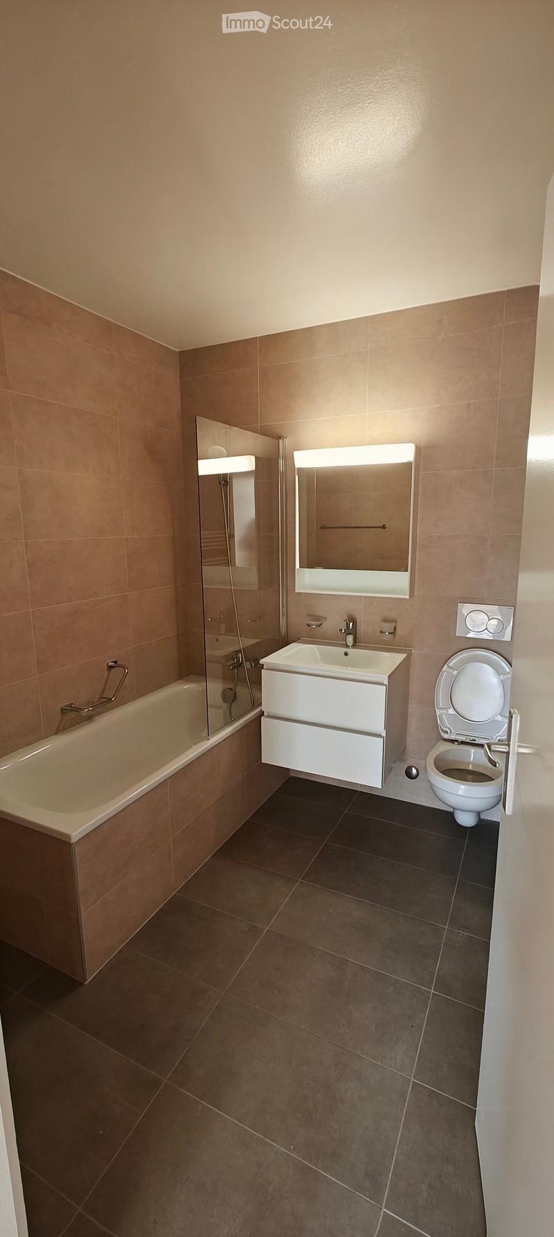 Modern bathroom with beige tiles, a bathtub, a sink with a vanity cabinet, a toilet, and a large mirror with built-in lighting.