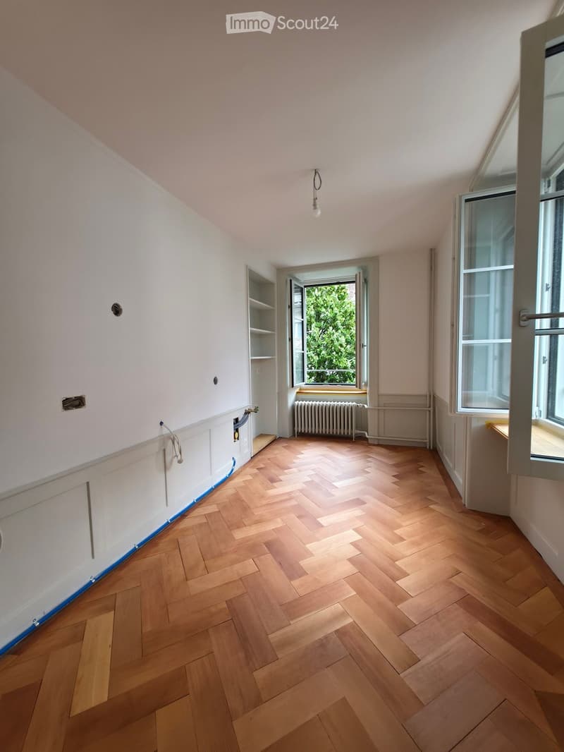 Empty room with wooden herringbone flooring, white walls, and large windows with open doors leading to a balcony. No furniture or equipment is present.