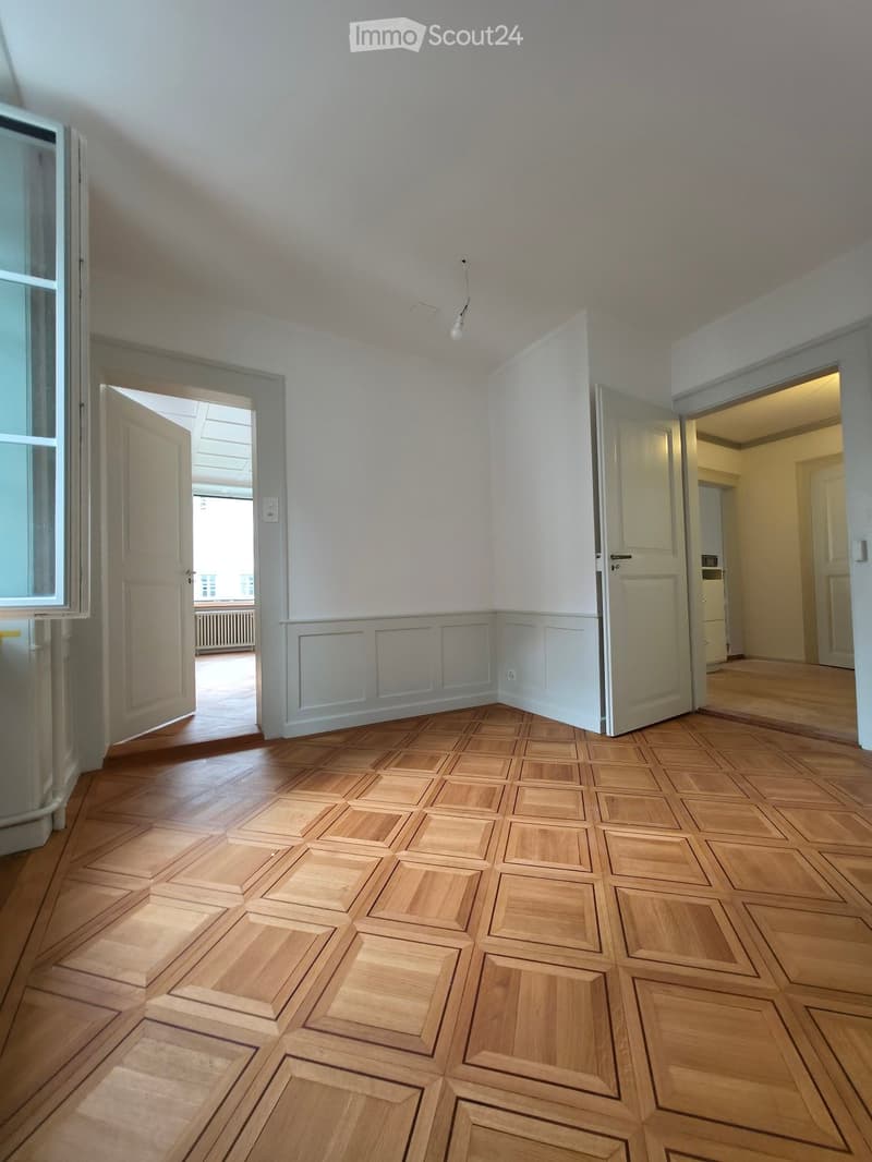 Empty room with wooden parquet flooring, white walls, and several doors leading to other spaces.