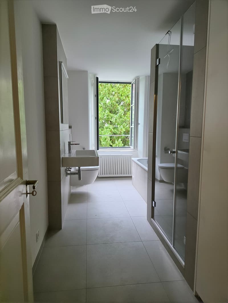 Modern bathroom with white tiles, a sink, toilet, bathtub, and a large window offering a view of greenery.