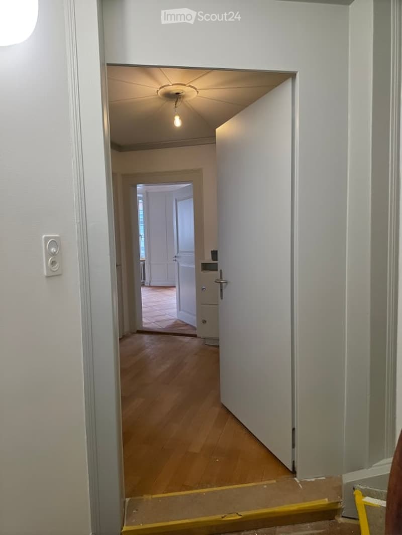 A narrow hallway with wooden flooring, white walls, and a ceiling light. An open door leads to another room with visible furniture and a tiled floor.