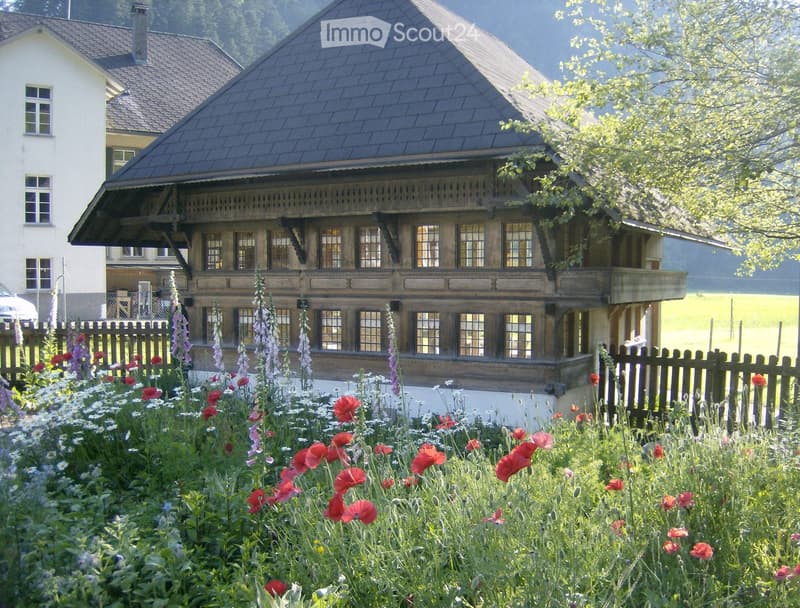 Traditional wooden house with a steep roof, large windows, and a balcony, surrounded by a garden with colorful flowers and a wooden fence.