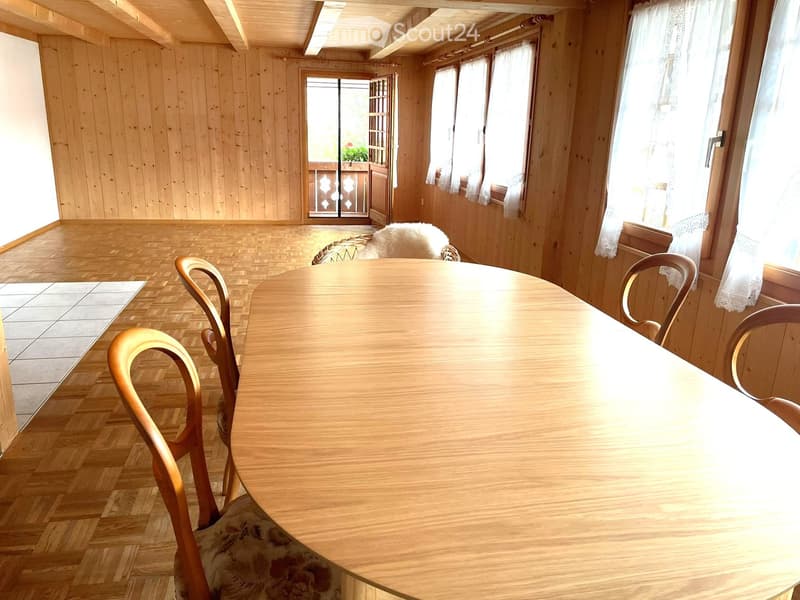 Bright wooden dining room with a round wooden table, four chairs, and white curtains on large windows. A doorway leads to a balcony with a small plant visible.