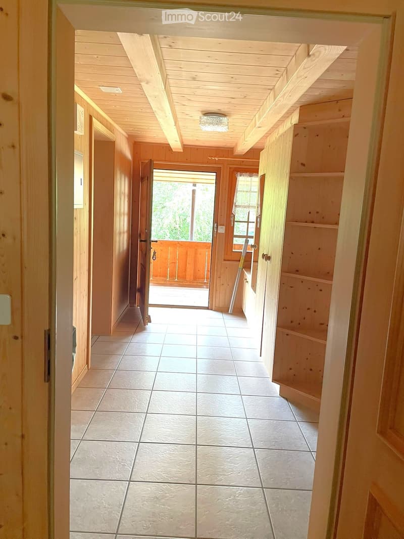 A long, wooden-paneled hallway with tiled flooring, leading to an open doorway that reveals a wooden deck and greenery outside. The hallway features built-in wooden shelving on one side and a ceiling light fixture.