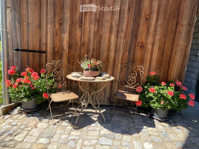 A small outdoor seating area with a round metal table, two ornate metal chairs, and potted flowers in terracotta pots, set against a wooden fence.