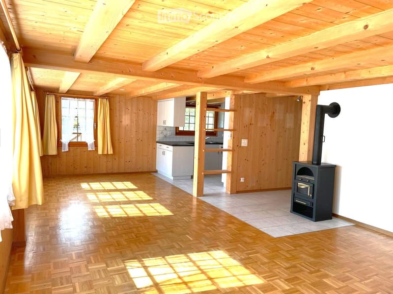 Bright, wooden-floored room with exposed wooden beams, a wood stove, and a small kitchen area with cabinets and a sink.