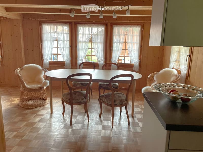 Dining room with wooden walls, herringbone parquet flooring, round wooden table surrounded by chairs, wicker chairs with fur cushions, and a glass bowl with fruit on a side table.