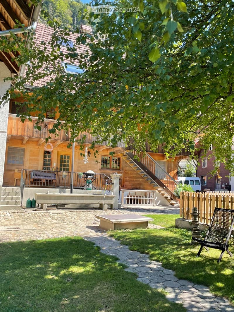 A well-maintained garden area with a stone-paved pathway, grassy lawn, wooden fence, and a wooden chair. The background features a wooden house with a balcony and a staircase leading to an upper level.