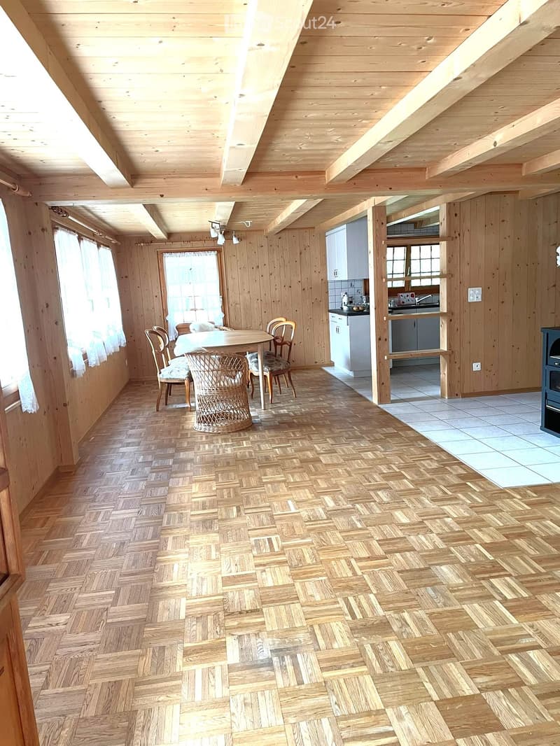 Open-plan living and dining area with wooden walls and herringbone parquet flooring, featuring a dining table with chairs, a wicker basket, and an adjacent kitchen area with cabinets and a sink.