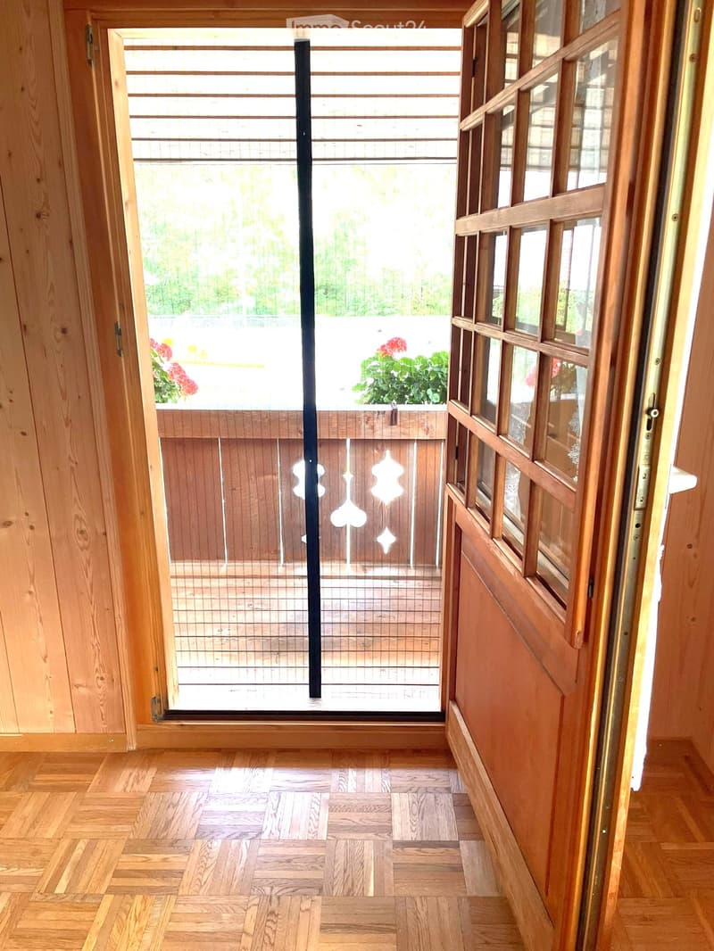 Wooden hallway with herringbone-patterned flooring, leading to a glass door opening onto a balcony with wooden railings and potted plants.