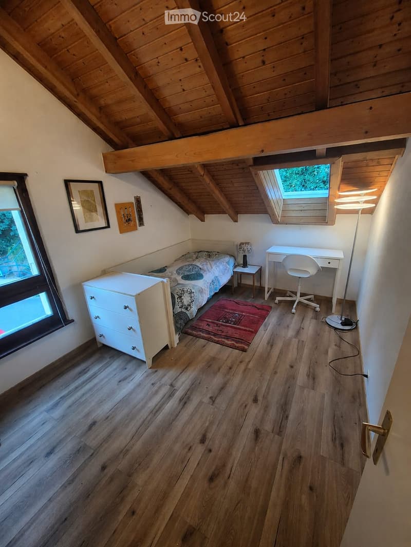 Cozy attic bedroom with wooden slanted ceilings, a single bed, a nightstand with a lamp, a small desk with a chair, a drawer unit, and a floor lamp. The room features wooden flooring and large windows with a view.