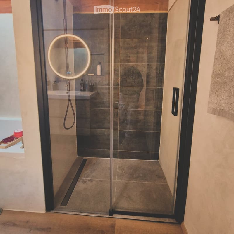 Modern bathroom with a glass-enclosed shower, a round LED mirror, and a towel hanging on the wall.