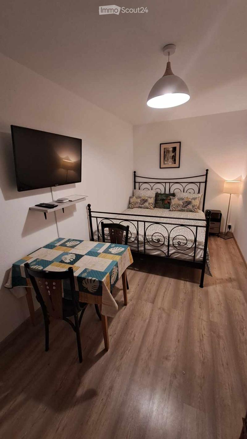 Cozy bedroom with a wooden floor, a metal-framed bed, a small table with a colorful tablecloth, a chair, a nightstand, a lamp, a picture frame, and a wall-mounted screen.