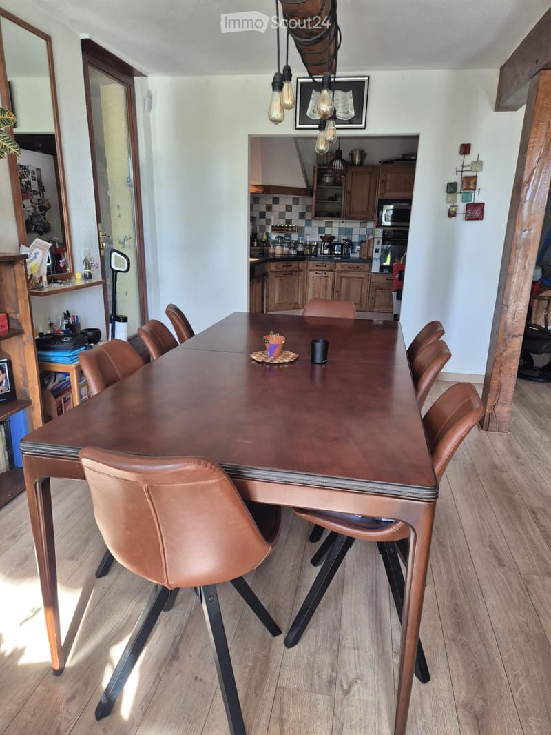 Dining room with a wooden dining table, brown leather chairs, a small potted plant, and a cup on the table. The room has wooden flooring and is connected to a kitchen area with wooden cabinets and appliances.