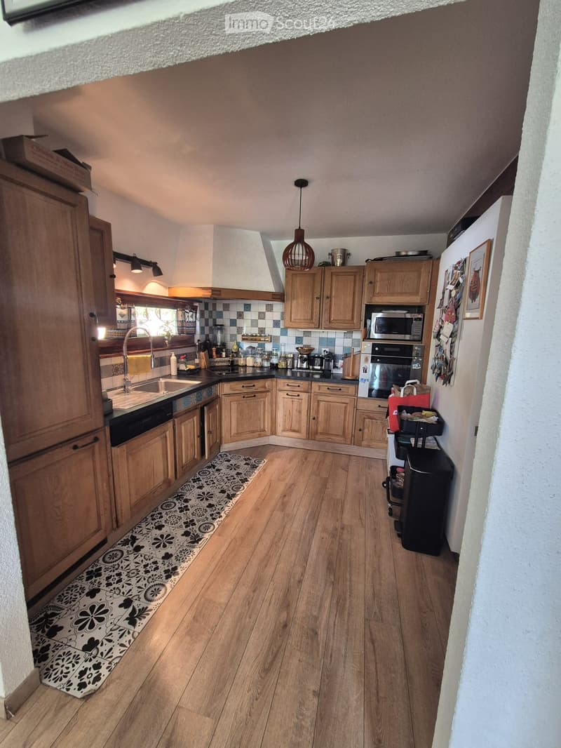 Well-lit kitchen with wooden cabinets, a sink, oven, microwave, and various appliances. The floor is wooden with a decorative black and white runner.