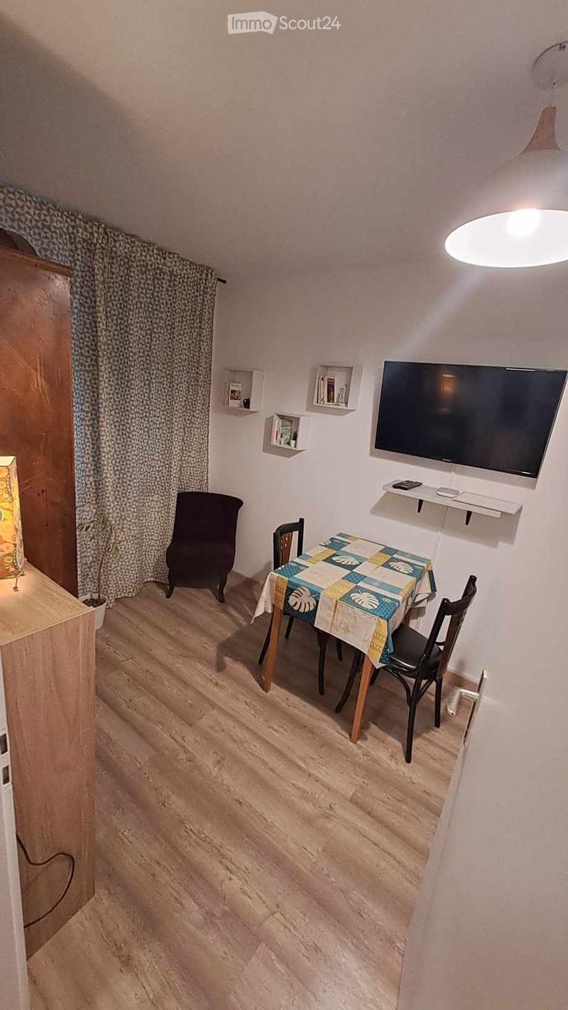 A small, cozy dining area with a wooden floor, a round table with a colorful tablecloth, four chairs, a mounted television, shelves, and a lamp.