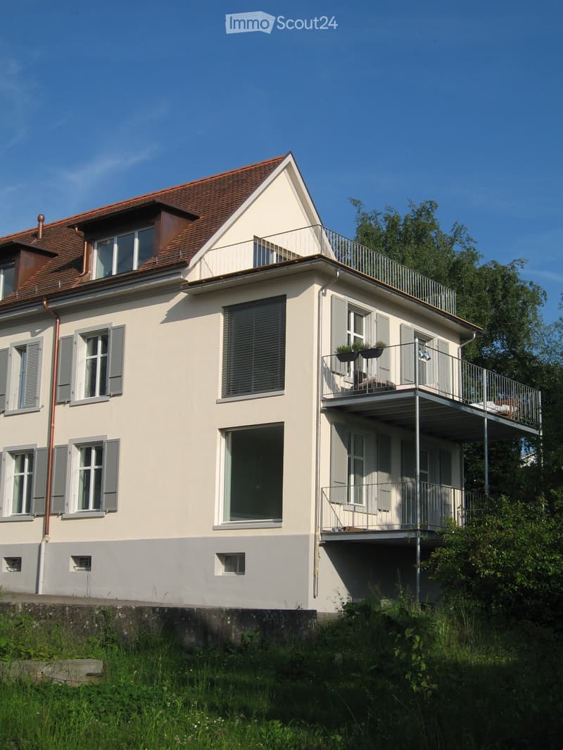 Two-story house with a balcony, grey shutters, and a brown tiled roof.