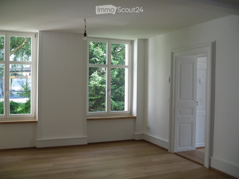 Empty room with wooden flooring, large windows, and a door leading to another space.