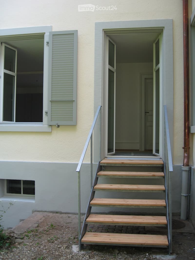 Exterior view of a building with a raised ground floor accessed by wooden steps with metal railings. The building has light-colored walls and multiple windows with shutters.