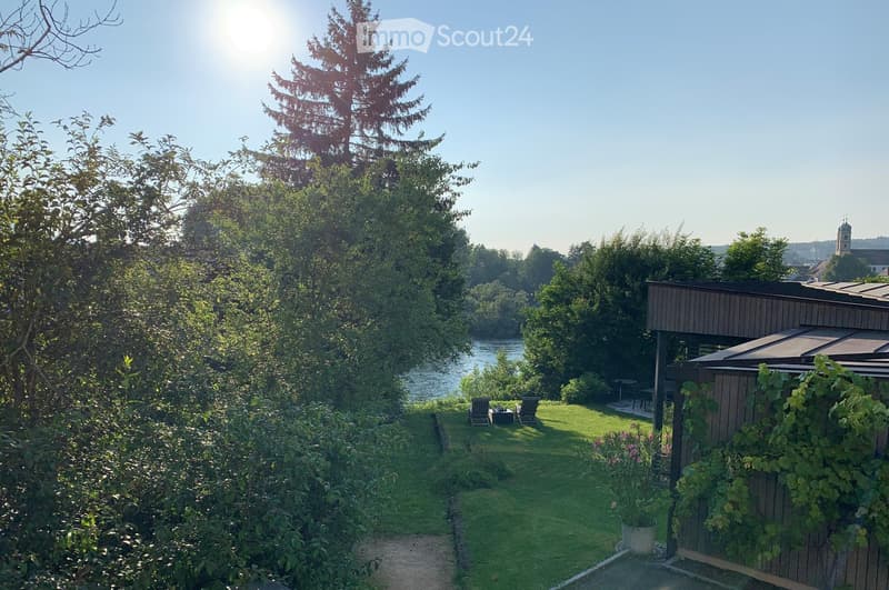 A serene garden area with a well-maintained lawn, a small wooden structure, outdoor seating, and a view of a river and distant town.