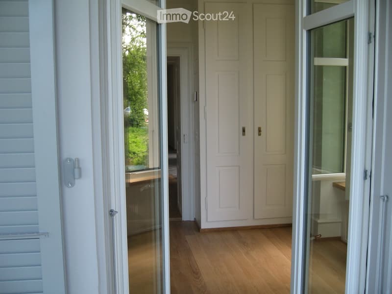 A well-lit hallway with wooden flooring, white walls, and several doors including a glass-paned door leading to another room.