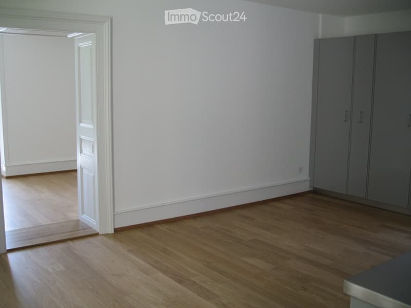 Empty room with light wooden flooring, white walls, and a built-in closet with grey doors. A doorway leads to another room.