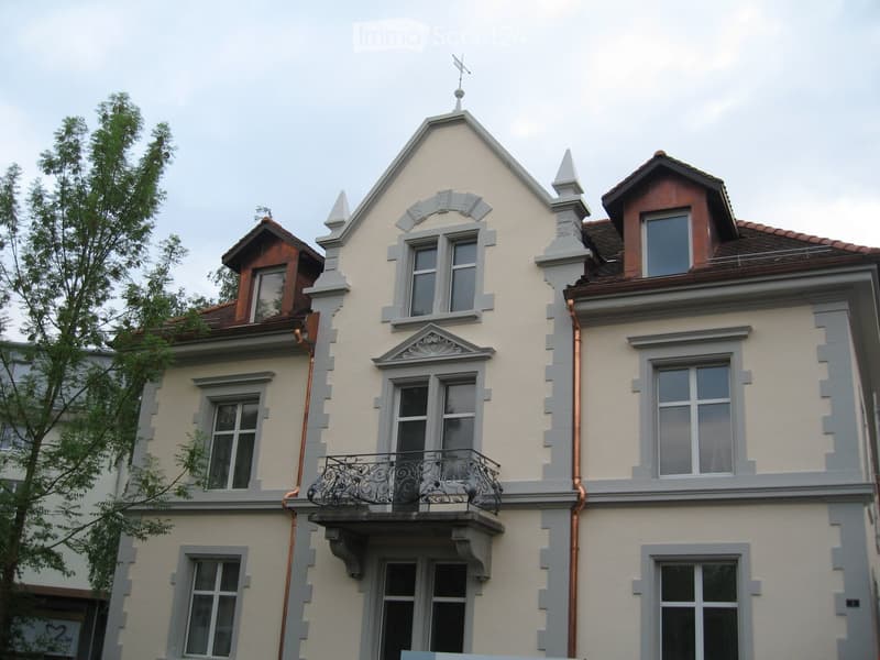 Two-story house with light yellow walls, gray trim, multiple windows, decorative roofline, and a small balcony with ornate railing.