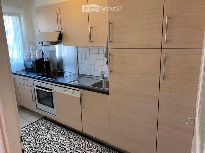 Modern kitchen with light wood cabinets, stainless steel appliances, and a black countertop. Features an oven, dishwasher, microwave, and a sink with a faucet. The floor has a patterned tile design.