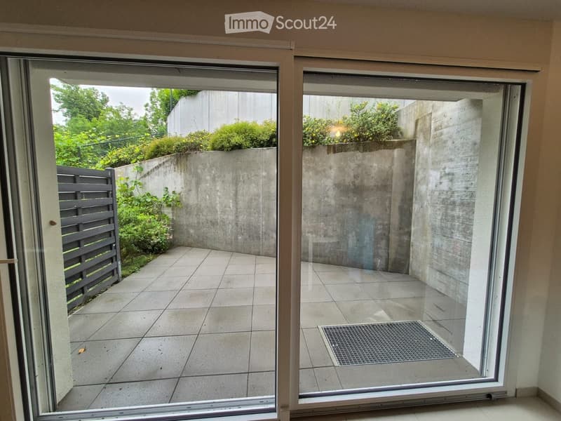 Two large sliding glass doors open to a tiled balcony area with a concrete wall, greenery, and a small metal grate on the floor.