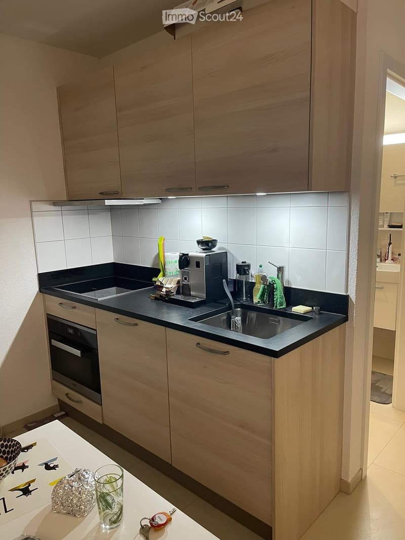 Modern kitchen with light wood cabinets, black countertops, stainless steel sink, and various appliances including a coffee machine and oven. A glass with a leaf design and keys are on the counter.