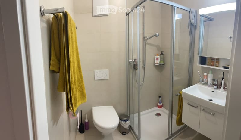 Modern bathroom with a toilet, sink, mirror, and glass-enclosed shower. Includes a yellow towel, toiletries, and a small trash can.