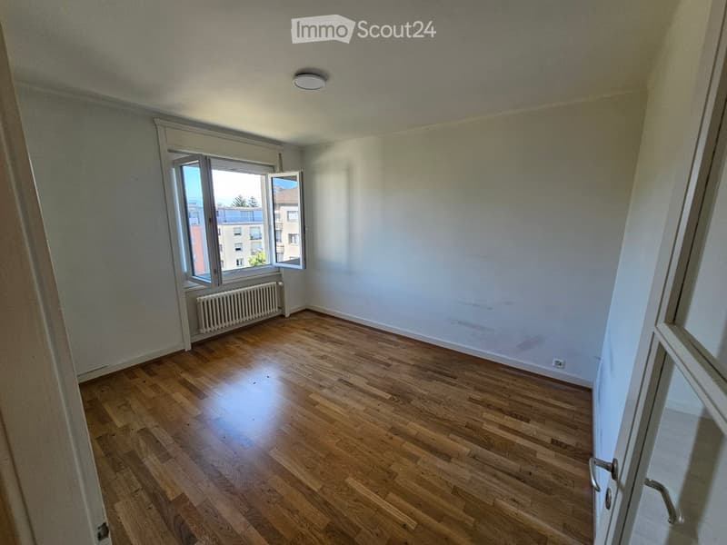 Empty room with wooden flooring, white walls, and a large window with a view of the outside. The room has a radiator under the window and a ceiling light fixture.