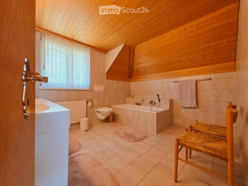 Bathroom with wooden ceiling, tiled walls, bathtub, toilet, sink, and a small wooden chair with woven seat.