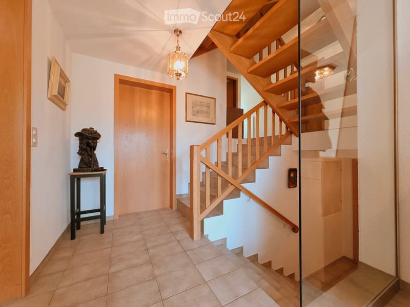Spacious hallway with wooden staircase, glass railing, pendant light, small table with sculpture, framed artwork, and tiled floor.