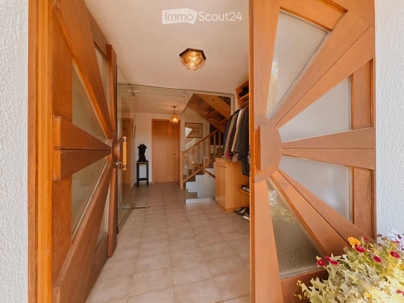 A well-lit hallway with wooden doors, a staircase leading upwards, and a small coat rack with jackets hanging on it. The floor is tiled, and the walls are painted in a neutral tone.