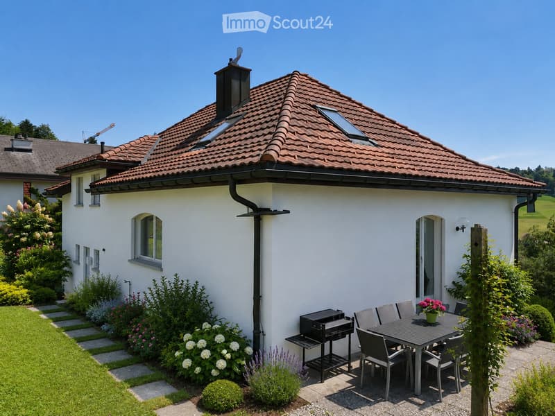 A modern house with a red-tiled roof and white walls, surrounded by a well-maintained garden. The patio features a dining table with chairs, a grill, and a vase of flowers. The garden includes neatly trimmed bushes, flowers, and a stone pathway leading to