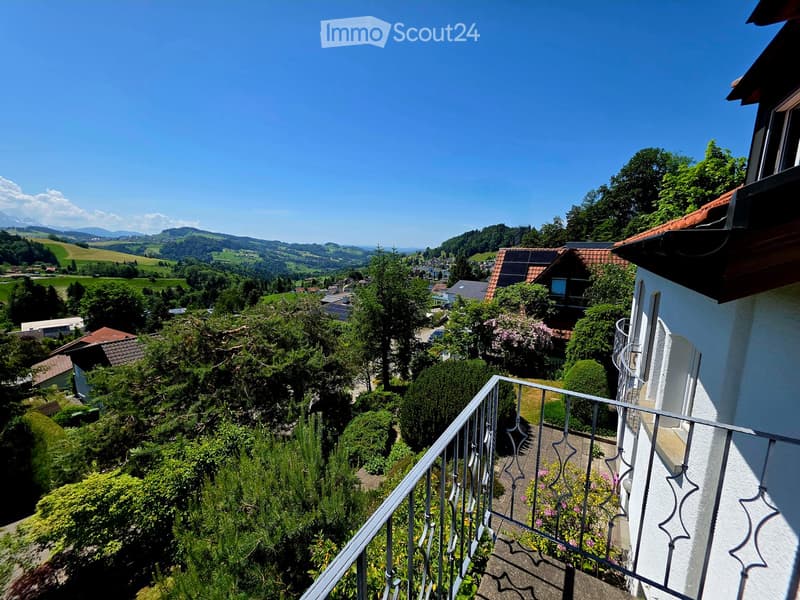 Balcony overlooking a scenic valley with houses, trees, and distant mountains under a clear blue sky.