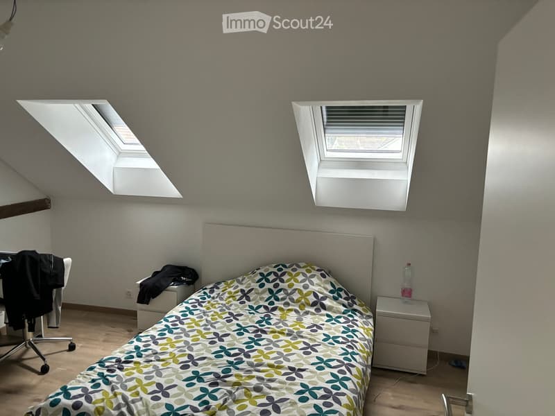 Bedroom with a bed, nightstand, chair, and a bottle on the nightstand. The room features skylight windows and wooden flooring.