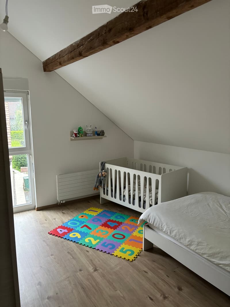 Bright, airy bedroom with wooden flooring, a single bed, a crib, a colorful foam mat with numbers, and a small shelf with toys.