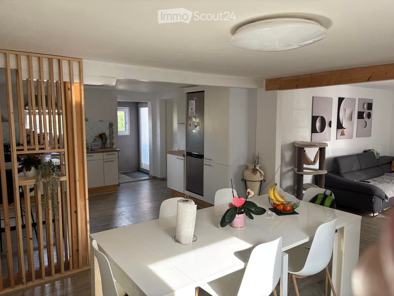 Modern living and dining area with a grey sofa, white dining table with chairs, cat tree, and a kitchen area with a refrigerator and dishwasher. The space features wooden flooring, white walls, and decorative artwork.