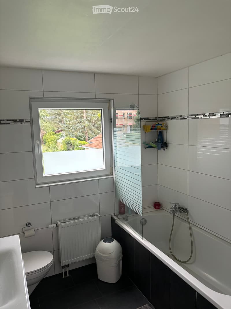 Modern bathroom with white tiles, a bathtub with a glass shower enclosure, a toilet, a radiator, and a small shelf with toiletries.