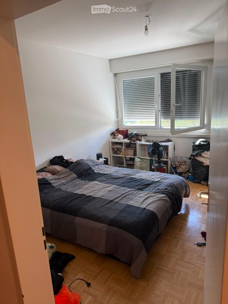 Bedroom with a large bed, bookshelf, fan, laptop, and various items scattered around. The room has wooden flooring and large windows with blinds.