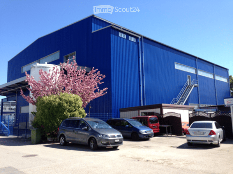 A large industrial-style building with a blue exterior and white trim, featuring a garage and parking area with several cars.