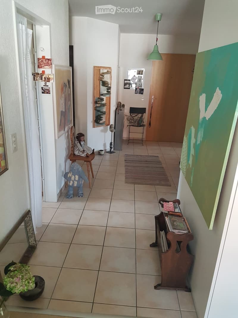 Hallway with tiled floor, white walls, and various decorations including a small chair with a doll, a shoe rack, a small table with books, and artwork on the walls.