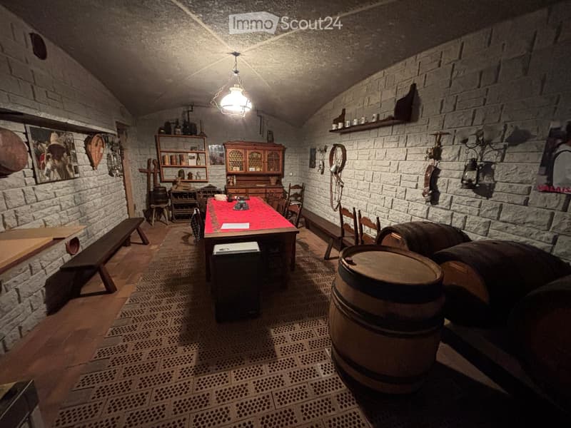 A rustic room with exposed brick walls, wooden floor, and a long wooden table with a red tablecloth. There are wooden benches and chairs, a wooden cabinet, barrels, shelves with various items, and a lantern hanging from the ceiling.