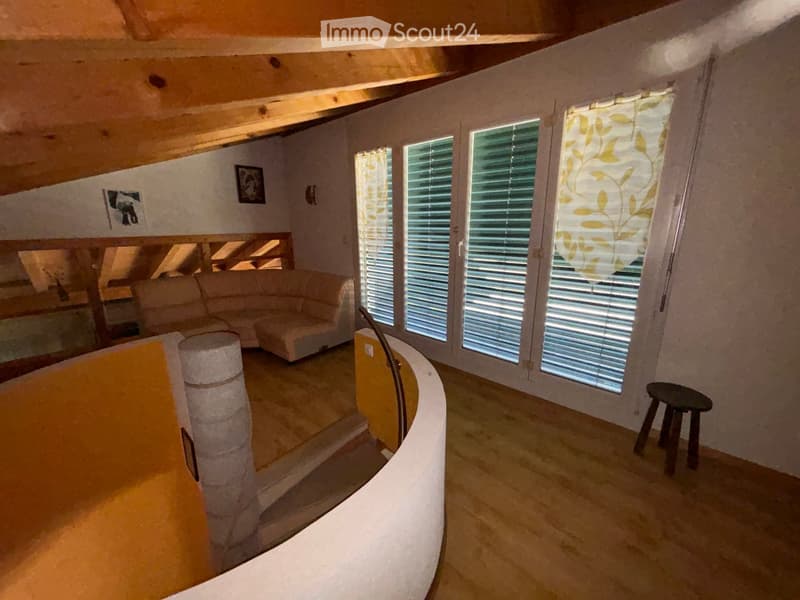 A cozy living room with a curved wall, large windows with blinds, a beige sofa, a small black stool, and a cat tree. The room has exposed wooden beams on the ceiling and a staircase leading upwards.
